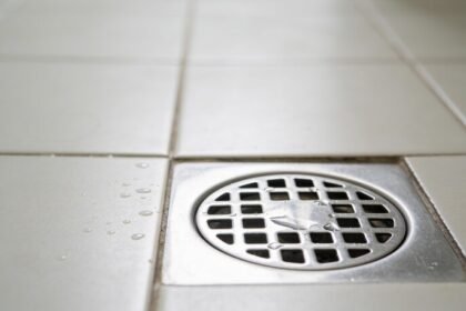A stainless steel floor waste drain set into pale tiles, photographed close to floor level with water beading on the tiles beside it.