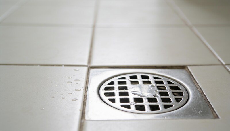 A stainless steel floor waste drain set into pale tiles, photographed close to floor level with water beading on the tiles beside it.