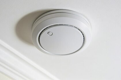 A plain round white smoke alarm mounted on a white ceiling, seen from below with soft daylight across it.