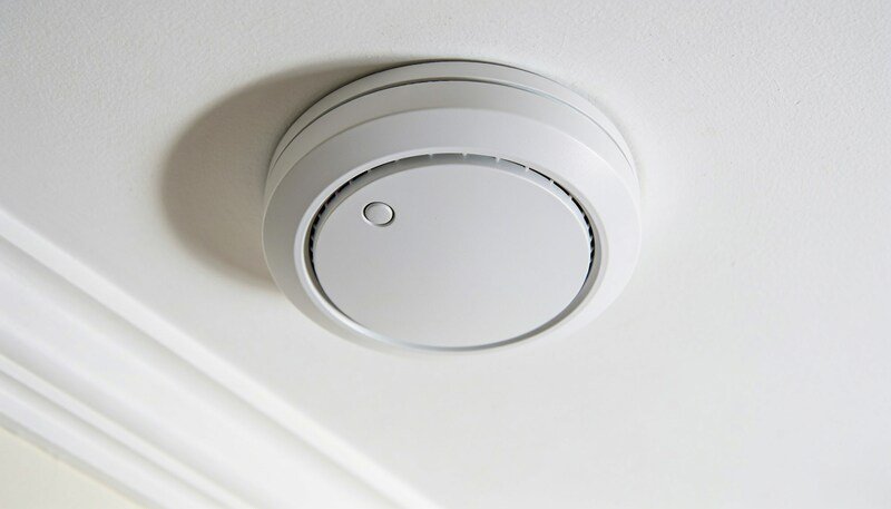 A plain round white smoke alarm mounted on a white ceiling, seen from below with soft daylight across it.