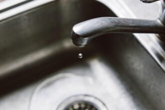 A plain brushed metal kitchen tap dripping into a stainless steel sink, seen close in with a single drop caught mid-fall.