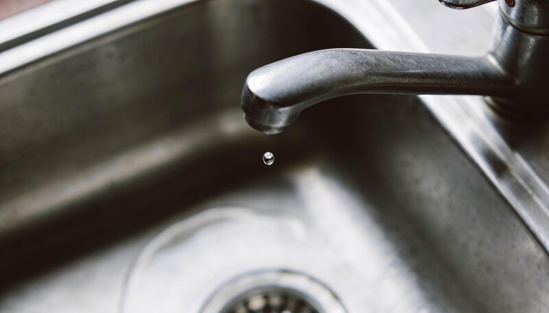 A plain brushed metal kitchen tap dripping into a stainless steel sink, seen close in with a single drop caught mid-fall.