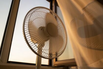 A plain white pedestal fan beside an open bedroom window at dusk, standing at the opening with a curtain moving beside it.