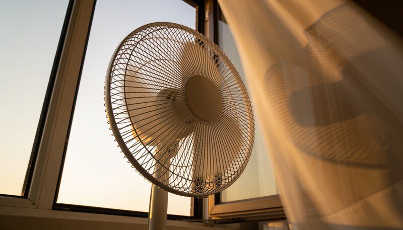 A plain white pedestal fan beside an open bedroom window at dusk, standing at the opening with a curtain moving beside it.