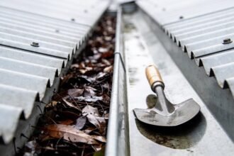 A metal roof gutter running between corrugated steel sheeting, seen close along its length, half packed with dark wet gum leaves and half cleared, with a trowel resting in the clear section.