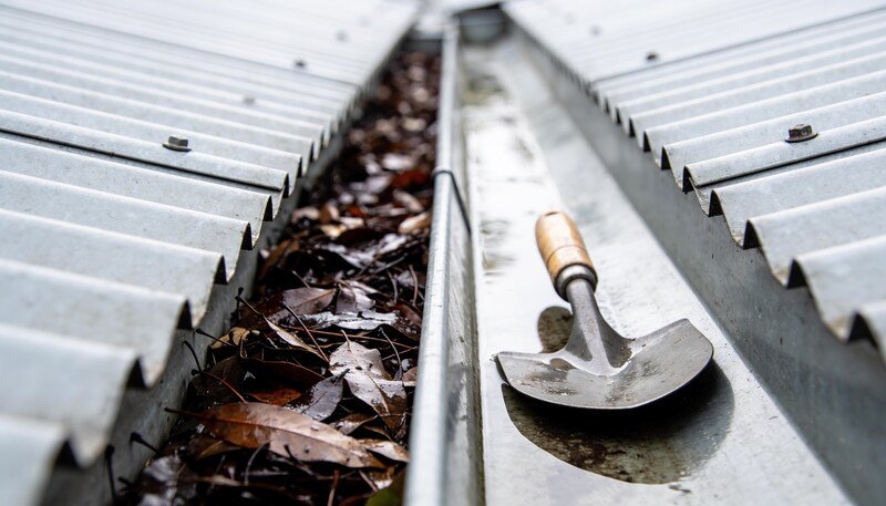 A metal roof gutter running between corrugated steel sheeting, seen close along its length, half packed with dark wet gum leaves and half cleared, with a trowel resting in the clear section.