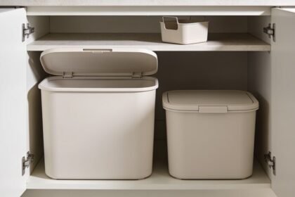 An open under-sink cupboard holding two plain bins of different sizes, the larger one open and the smaller one lidded, with a small caddy on the shelf above.