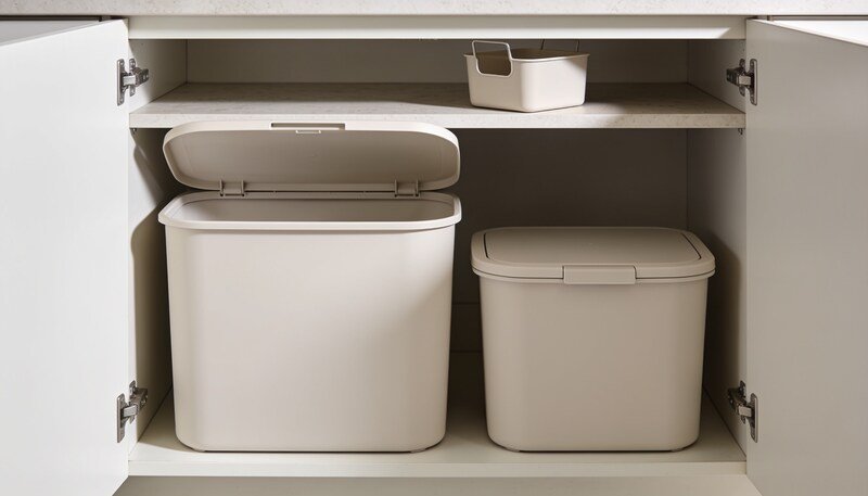 An open under-sink cupboard holding two plain bins of different sizes, the larger one open and the smaller one lidded, with a small caddy on the shelf above.