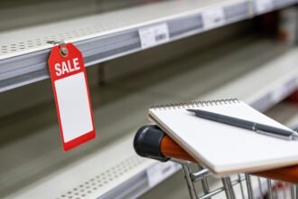 A blank sale ticket on an empty supermarket shelf edge beside a shopper's notepad on a trolley handle.