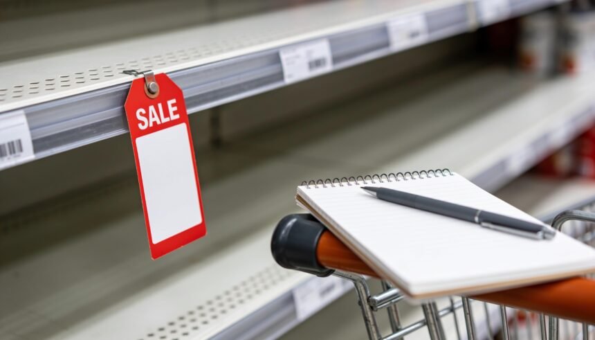 A blank sale ticket on an empty supermarket shelf edge beside a shopper's notepad on a trolley handle.