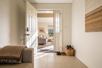 A tidy entryway and lounge seen through an open front door, shoes in a basket and a folded blanket on the sofa.