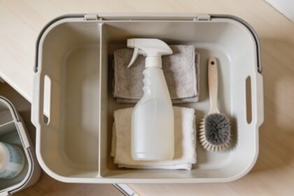 Looking down into a plain cleaning caddy on a kitchen bench holding a spray bottle, folded microfibre cloths and a scrubbing brush, with a second smaller caddy at the edge of frame.