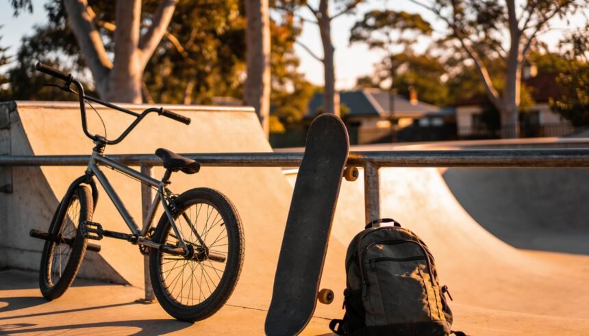 A skateboard and bike leaning against a skate park rail in late afternoon light, backpack on the ground beside them.