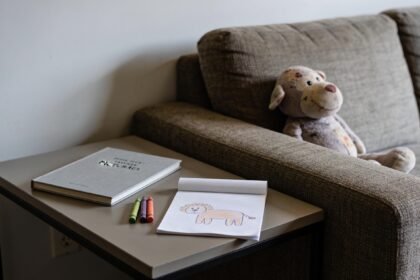 A picture book, folded drawing and crayons on a side table beside a sofa with a soft toy.
