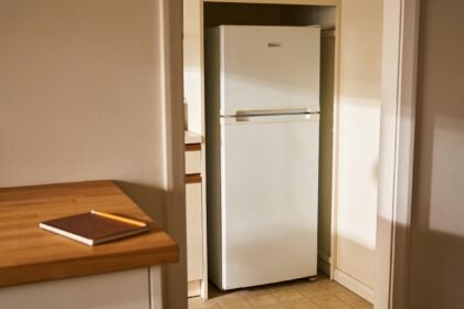 A refrigerator with space around it beside a kitchen bench holding a notebook and pencil.