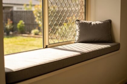 An empty padded window perch on a sunny windowsill with a flyscreen visible behind the glass.