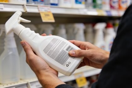 Hands holding a cleaning spray bottle with its rear label facing the shopper in a supermarket aisle.