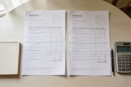 Two payroll documents beside a notebook, pen and calculator on a dining table.