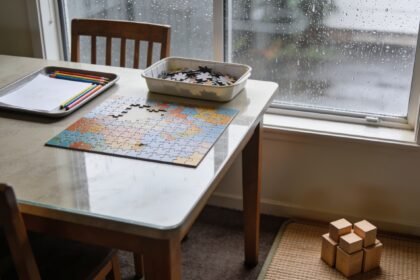 Drawing supplies and a partly finished puzzle arranged on a shared table beside a rainy window.