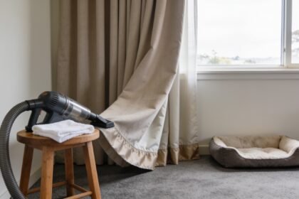 Plain curtains and carpet beside a moved pet bed, with a vacuum attachment and white cloth on a stool.